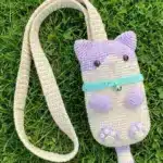 The finished purple and beige crochet cat phone bag with its long strap lying flat on green grass